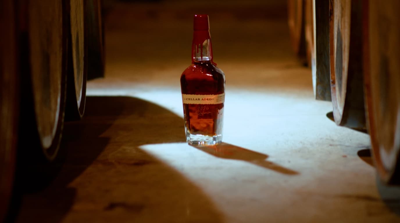 Maker's Mark Cellar Aged 2026 Release bottle with red wax seal spotlit on a limestone cellar floor between oak barrels
