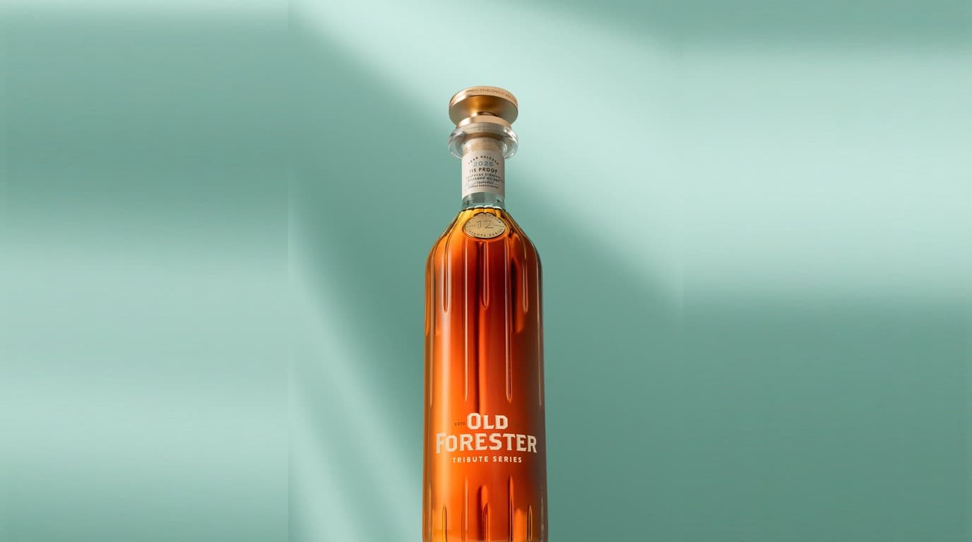 The 2026 Old Forester Tribute Series bottle, a fluted amber decanter with a gold metallic stopper, a 12 Year medallion at the shoulder and a 115 proof neck label, photographed against a pale green studio backdrop
