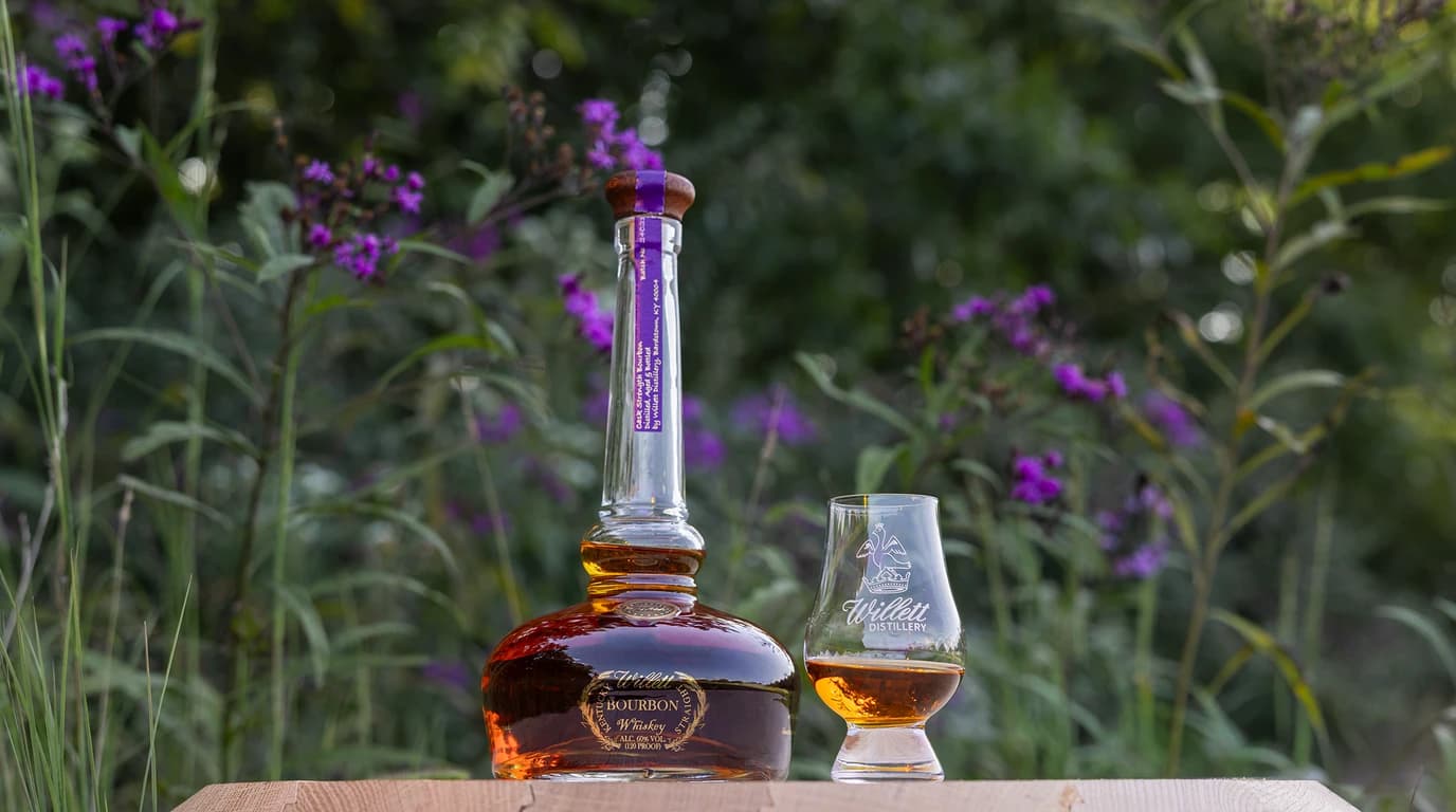 A bottle of Willett Pot Still Reserve Cask Strength bourbon, purple neck strip reading Cask Strength Bourbon, beside a Willett Distillery Glencairn glass on a wooden plank in front of blooming ironweed